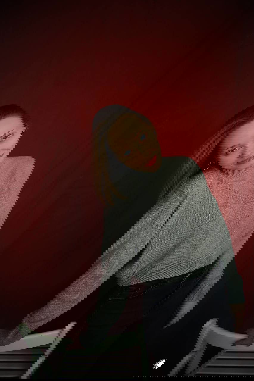 Портрет женщины на красном фоне: светлая кожа, серый свитер, человек опирается на белый стул, спокойное выражение лица.