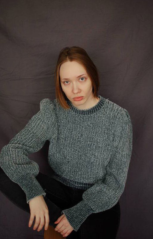 Портрет женщины в сером вязаном свитере на нейтральном фоне, сидит с опорой на ногу, спокойный взгляд в камеру.