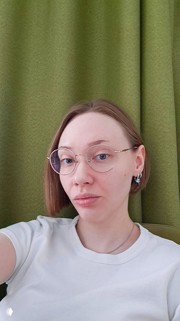 Портретное селфи женщины в круглых очках на фоне зелёных штор, нейтральное выражение лица, верхняя часть корпуса в кадре.