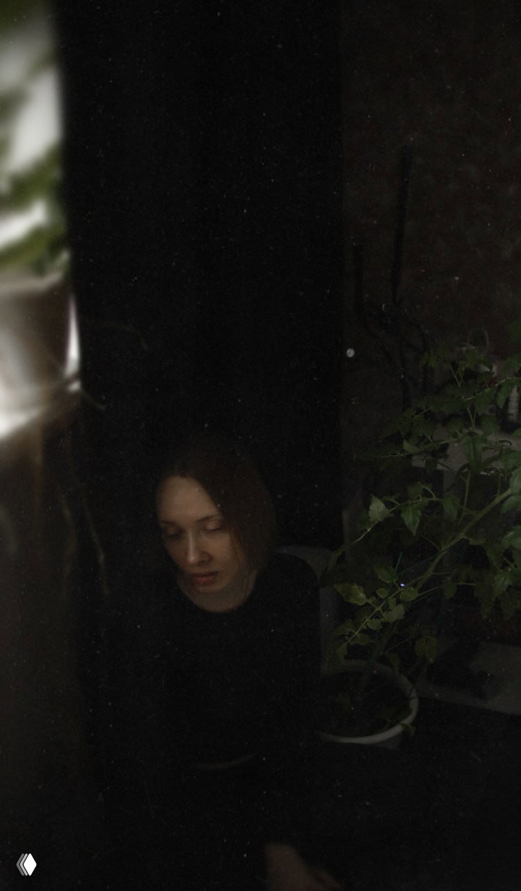 Портрет женщины в полутьме у окна и домашних растений: задумчивое лицо, мягкий свет и интерьерная сцена для астрологического поста