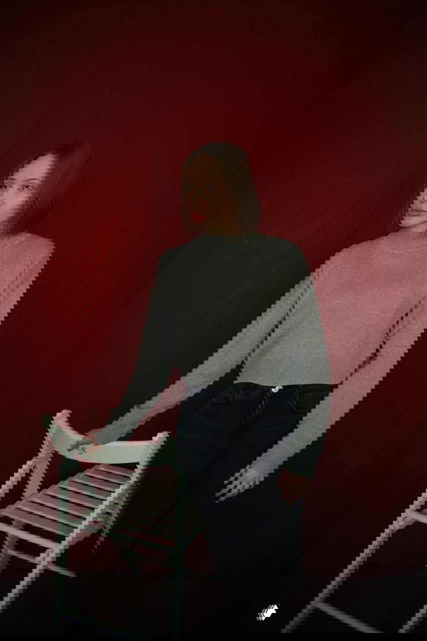 Фотография женщины в сером свитере на красном фоне, стоящая у стула — нейтральный портрет, подходящий для авторского астроблога.