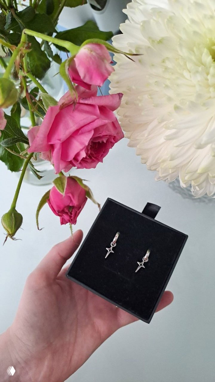 Рука держит чёрную подарочную коробочку с маленькими серьгами в форме звёзд; на заднем плане розовые розы и крупный белый цветок на светлом столе.