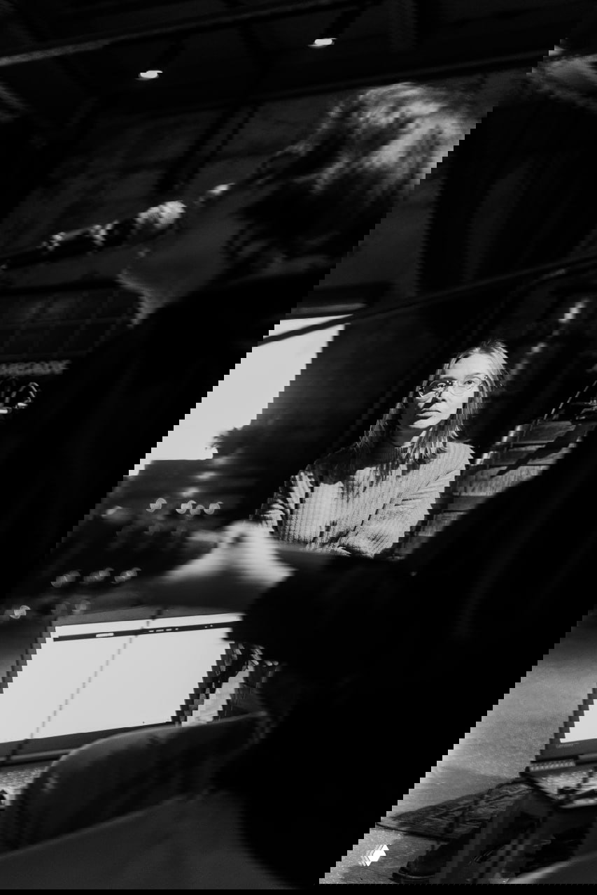 Чёрно‑белая фотография репетиции: музыкант играет на гитаре и поёт в микрофон, на заднем плане слушатель и открытый ноутбук — сцена репетиции.