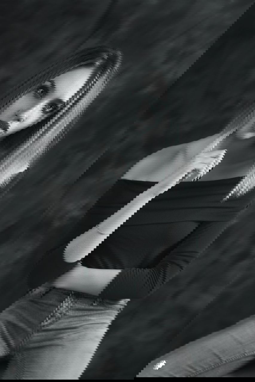 Чёрно-белый студийный портрет молодой женщины в джинсах и с открытыми плечами, задумчивый взгляд, нейтральный фон, спокойная поза.