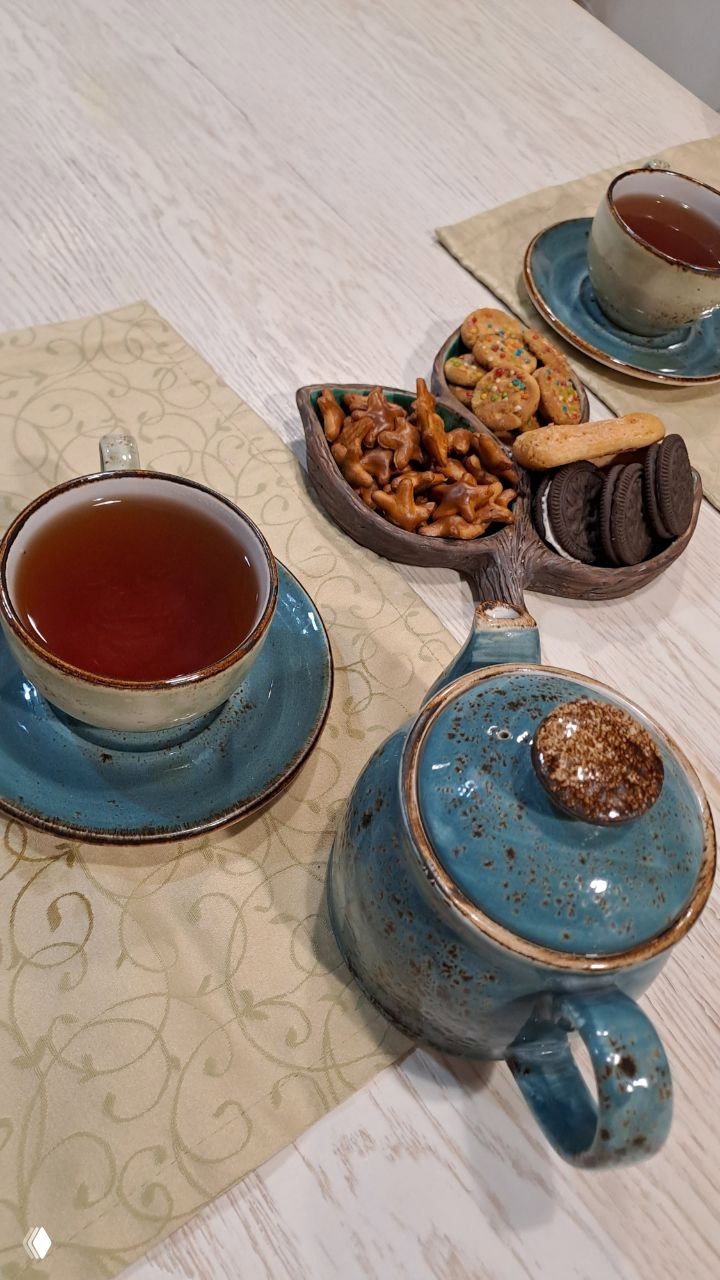 Чайный столик с керамическим чайником и чашками на салфетке, рядом небольшая тарелка с печеньем, кренделями и орешками — уютная сервировка.