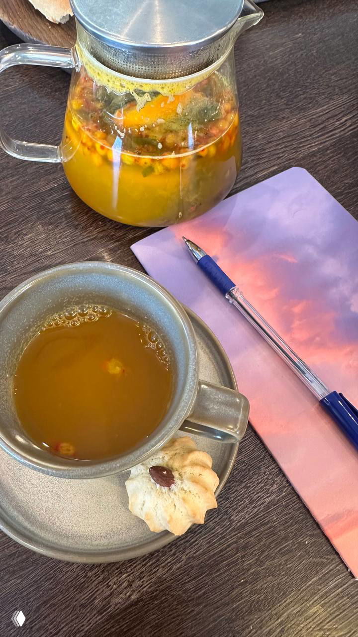 Чай в чашке и прозрачный заварник на столе рядом с фиолетовым блокнотом и ручкой, небольшое печенье — рабочая атмосфера подготовки.