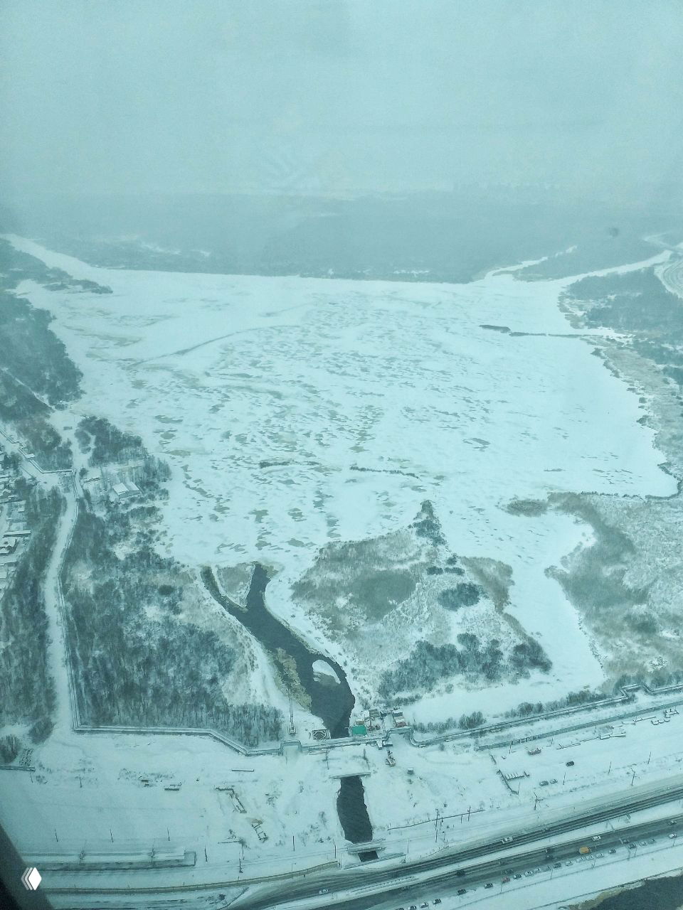 Широкая панорама замёрзшего залива и прибрежных ландшафтов, снег, лёд и редкие темные протоки воды на фоне далёкого горизонта.