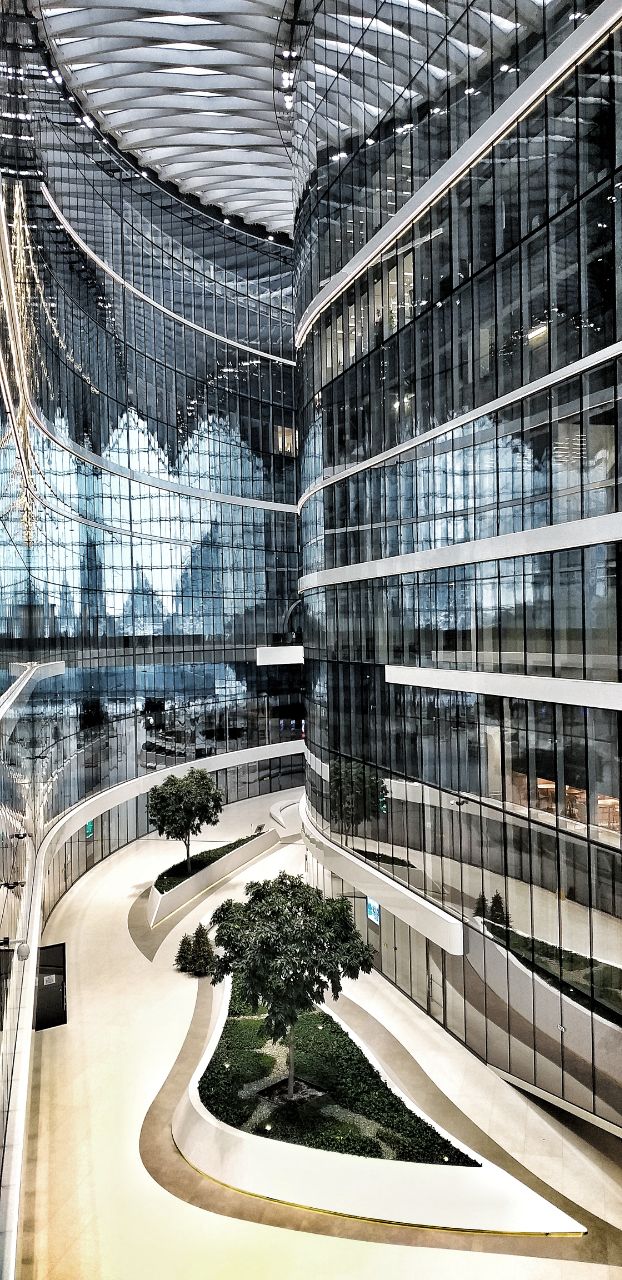 Внутренний простор атриума Лахта‑центра с изогнутыми стеклянными фасадами, отражениями и растениями в светлом современном пространстве.