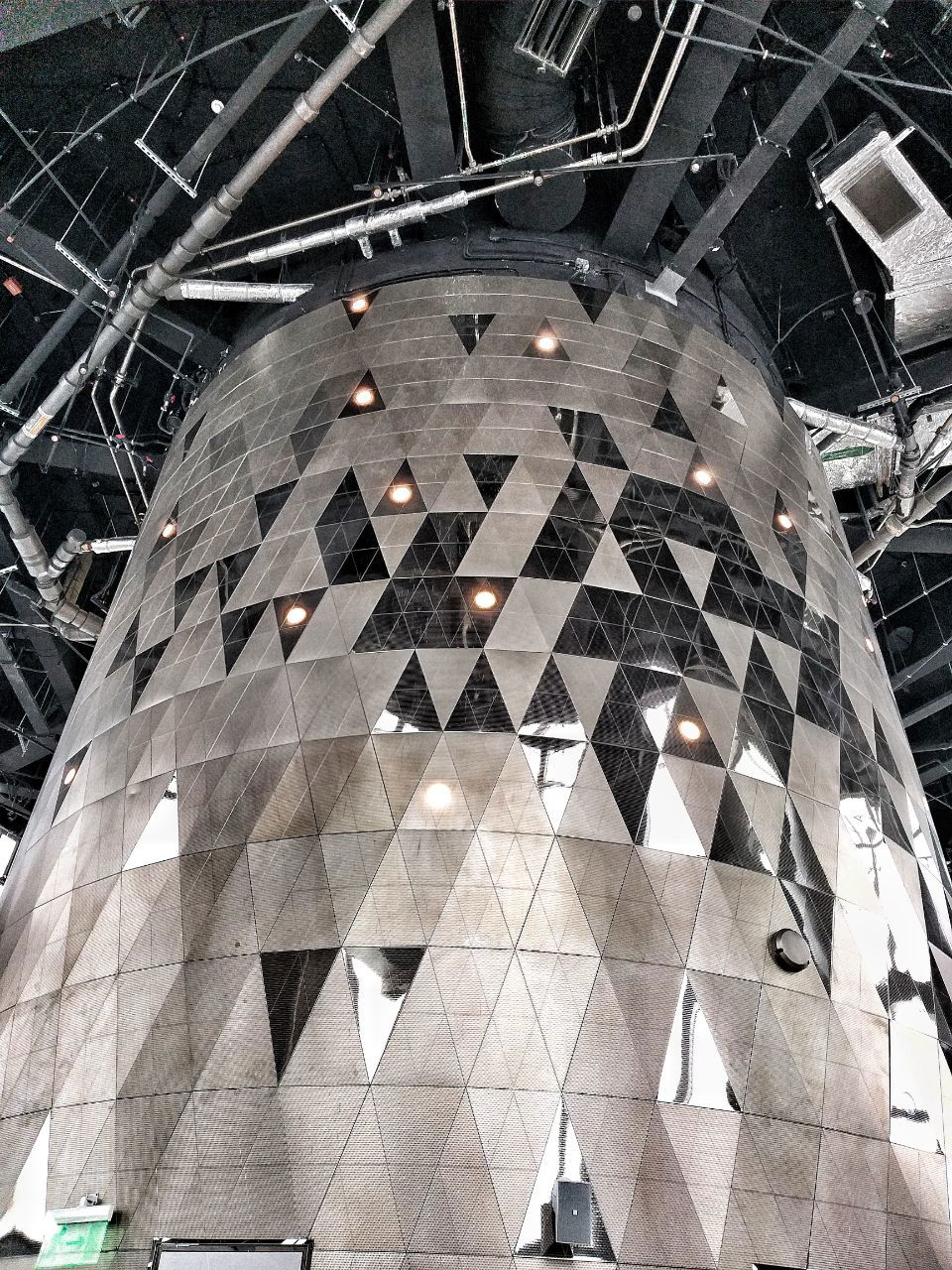 Крупный план цилиндрической декоративной колонны с фасеточной мозаикой из треугольников из металла и зеркал в интерьере Лахта‑центра.