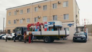 Праздничная платформа на грузовике с моделями зданий и людьми на фоне административного здания во время подготовки к шествию в Благовещенске