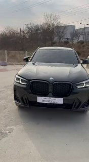 Небольшое вертикальное фото фасада автомобиля спереди; чёрный BMW X4 на ровной площадке, видны дневные фонари и решётка радиатора.