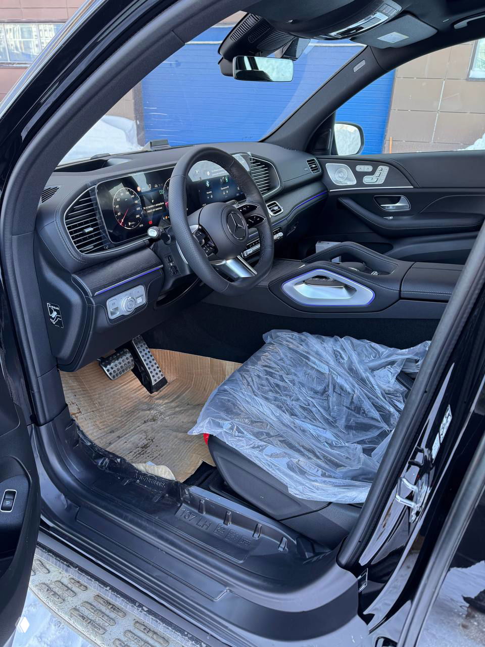 Водительская зона GLS 450d: вид на руль, мультимедийный дисплей и переднюю панель в дневном освещении, новые пластиковые чехлы на сиденьях.