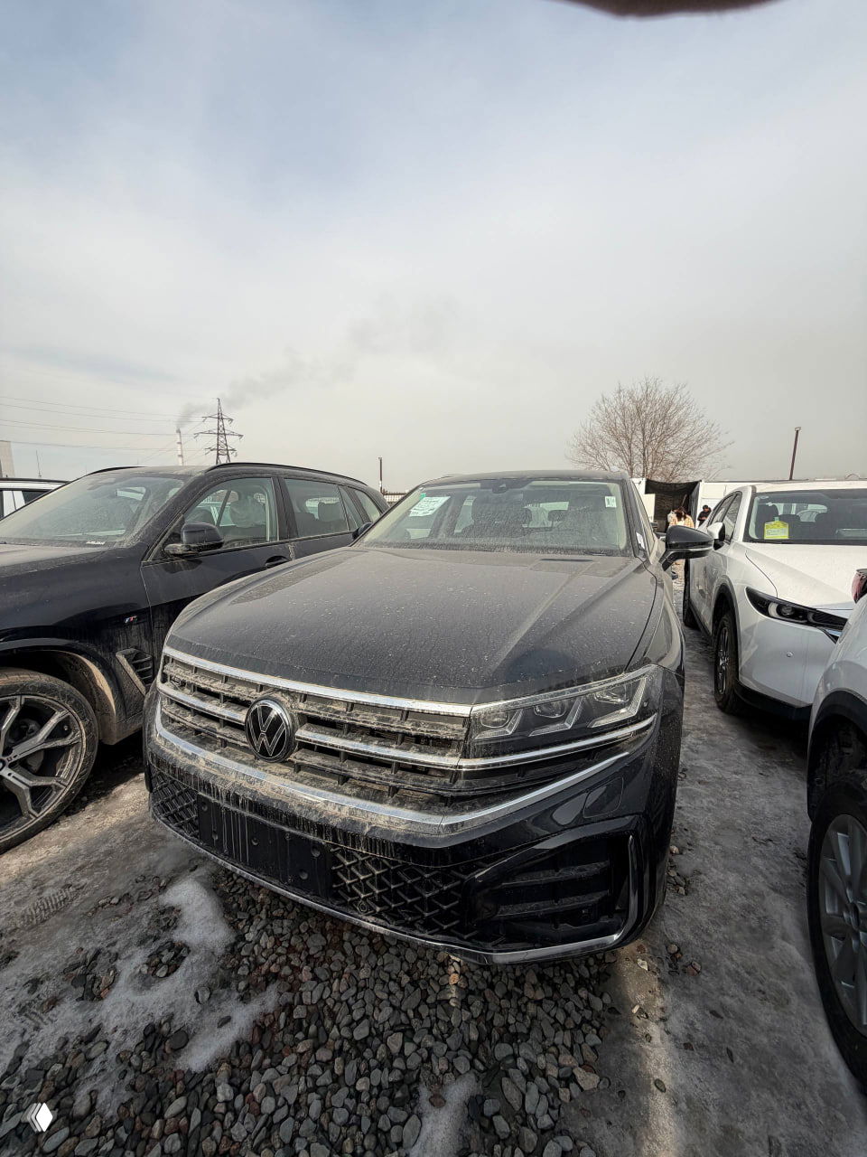 Вид спереди на чёрный Volkswagen Touareg, автомобиль припаркован между другими машинами на гравийной площадке с наледью.