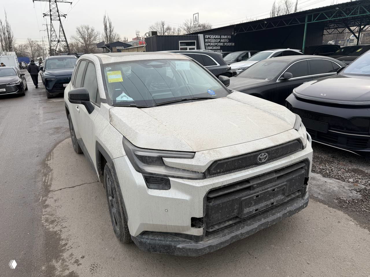 Крупный вид спереди на Toyota RAV4 6‑поколения: фары, бампер и грязь на кузове, автомобиль стоит возле других машин на площадке.