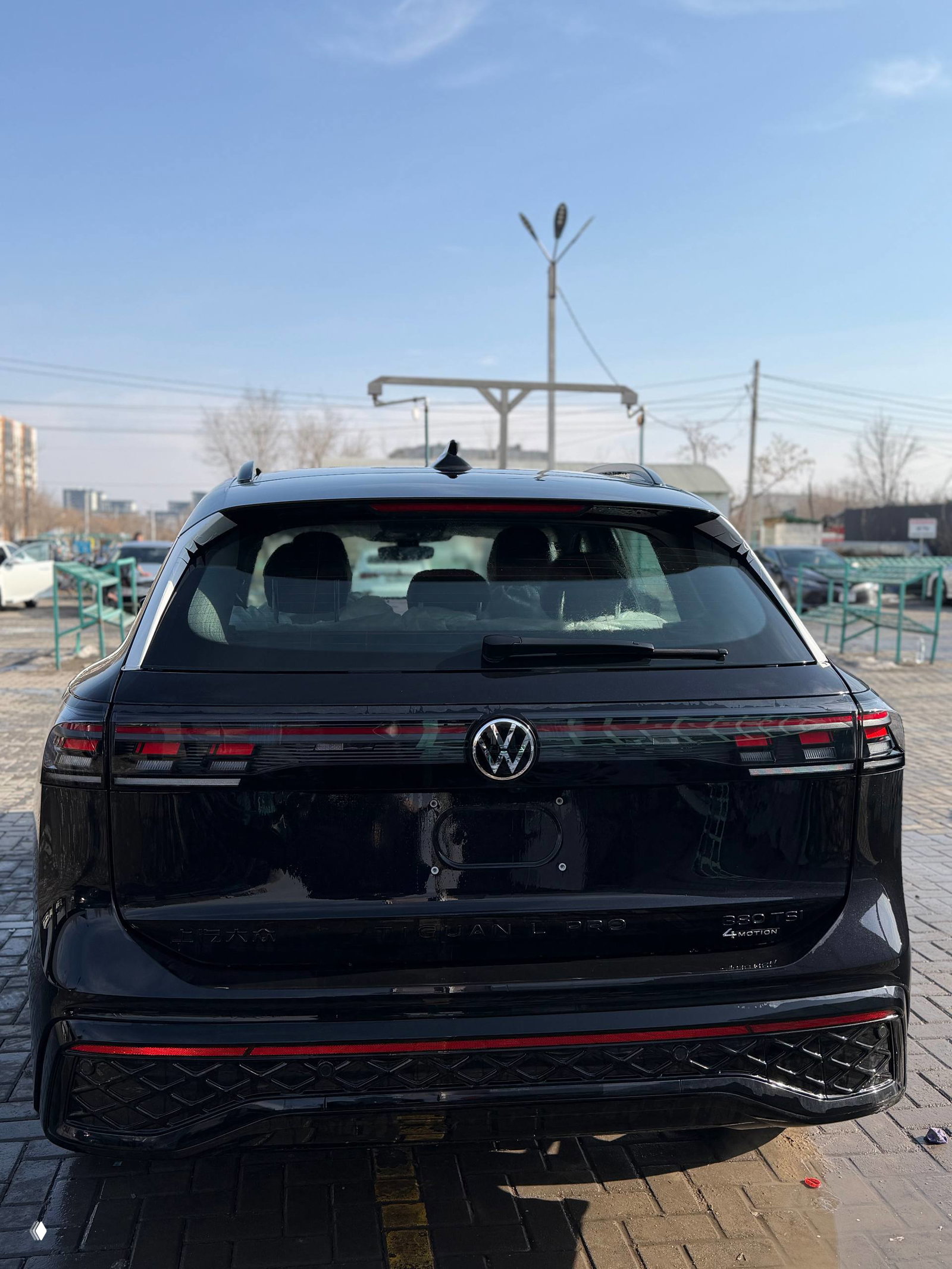 Задняя часть Volkswagen Tiguan L Pro: светодиодная полоска стоп-сигналов, эмблема VW и багажная дверь на фоне стоянки.