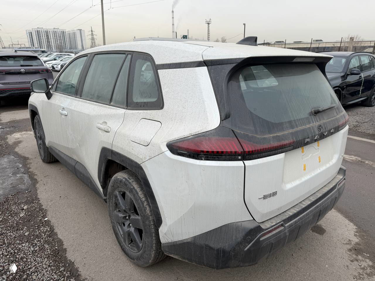 Трёхчетвертной вид сзади слева на Toyota RAV4 6‑поколения, видно задние фонари, колёса и общий силуэт автомобиля на площадке.