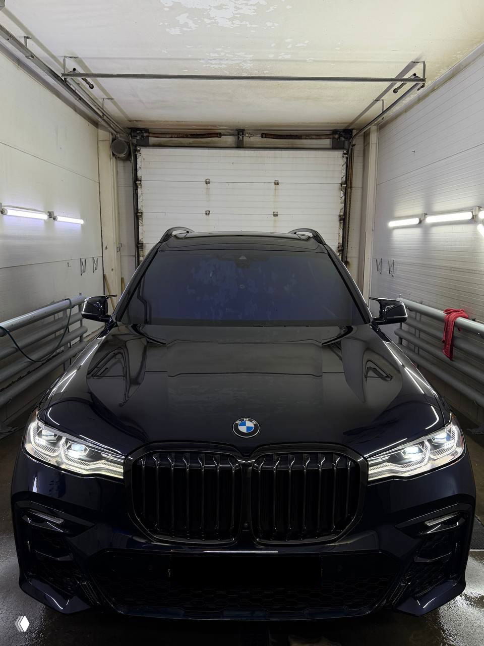 BMW X7 тёмного цвета снят спереди в гаражной мойке: вид капота, решётки, поток света отражается на лаке, автомобиль в родной краске и в пленке.