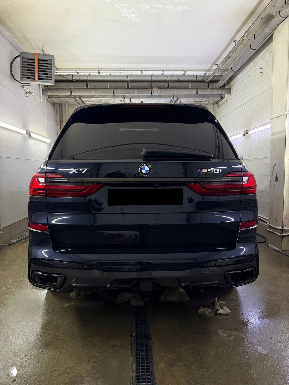 Задняя часть BMW X7 с модельным шильдиком X7 и M50i, видно двойные выхлопные патрубки и прицепной фаркоп, номер затемнён.