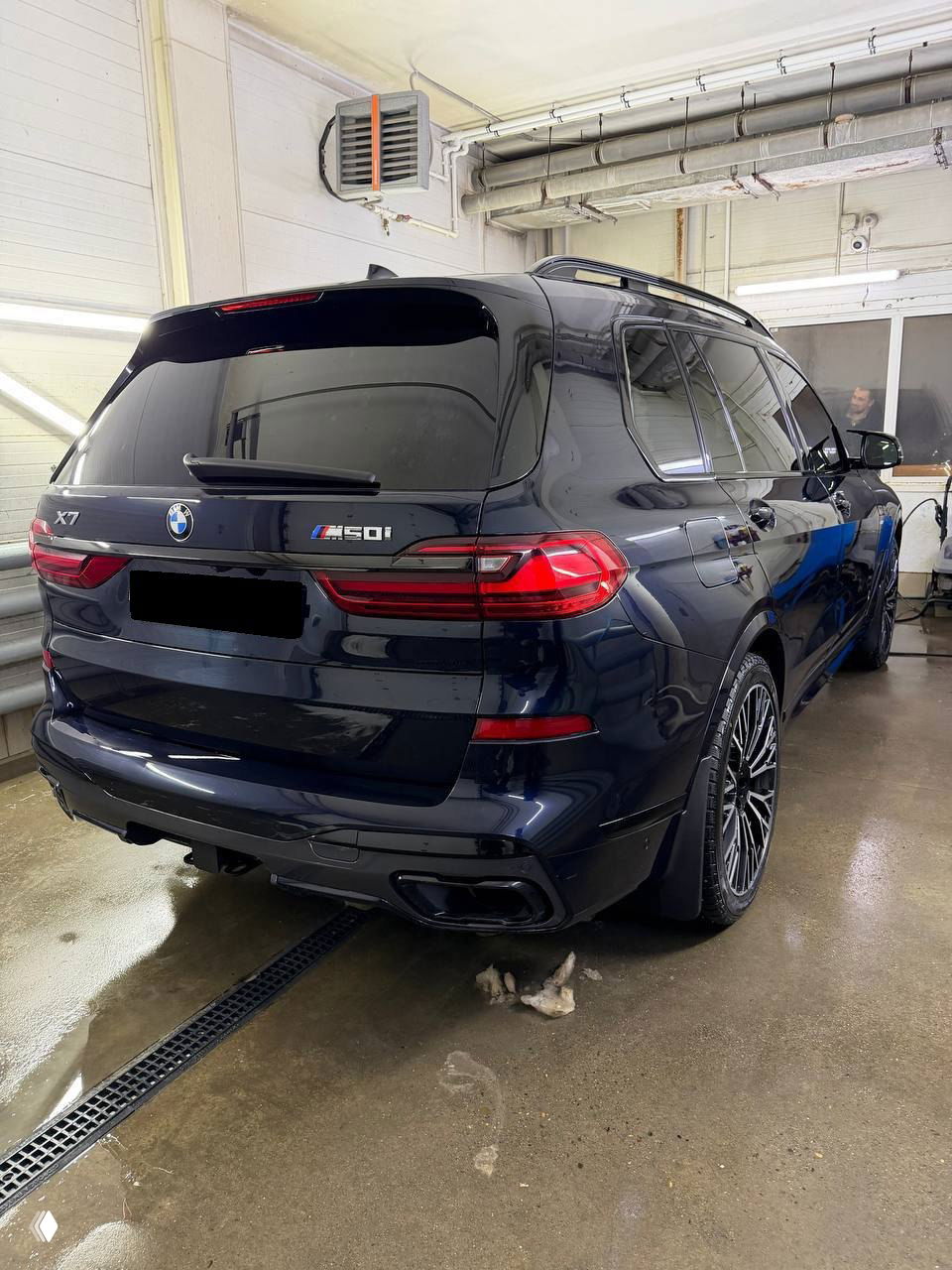 Угол сзади справа BMW X7 в моечном боксе: профиль, разноширокие колёса и боковые линии кузова, видна обшивка и отражения в лаке.