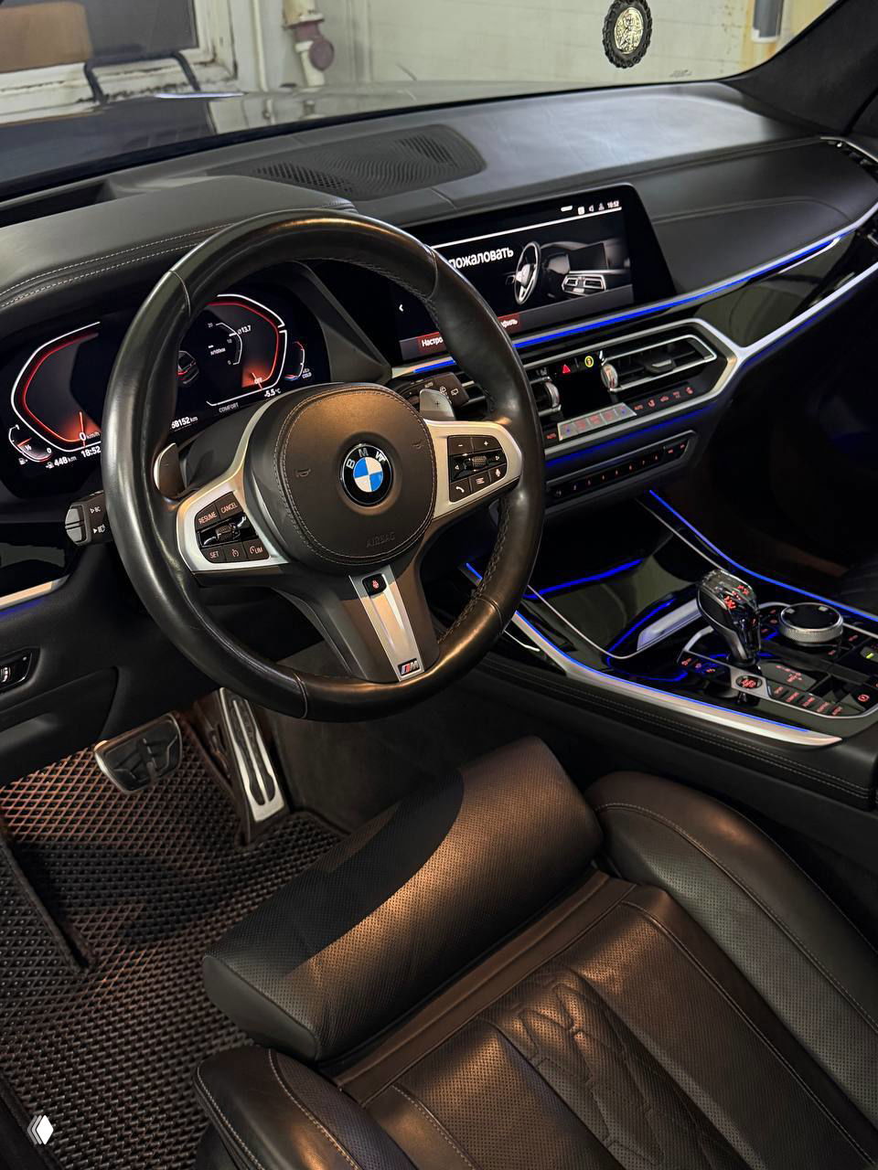 Салон BMW X7: вид на руль M, центральную консоль с подсветкой, большие дисплеи и элементы управления, отделка кожа и алюминий.