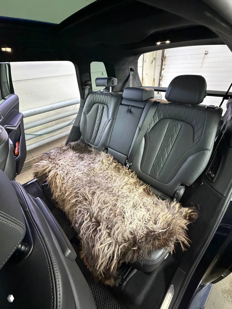 Задний ряд салона BMW X7 с кожаными сиденьями и декоративной накидкой на сиденье, видна отделка дверей и потолок алькантара с подсветкой.