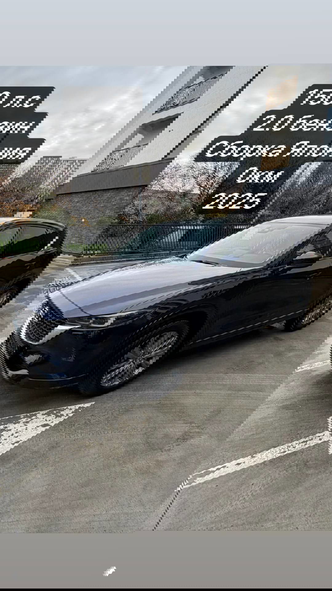 Тёмно‑синяя Mazda CX-5 2025 на открытой парковке; снимок показывает переднюю часть и фары, на изображении текстовые стикеры с характеристиками и нулевым пробегом.