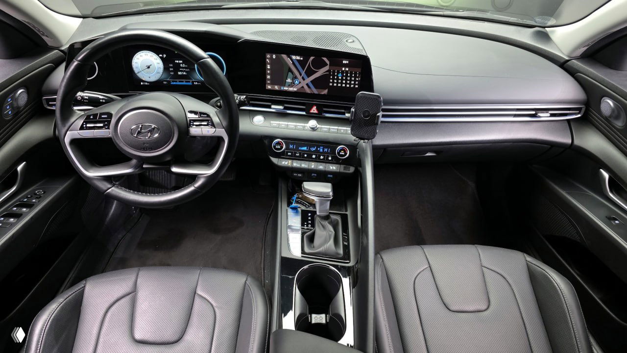 Панорамный кадр салона Hyundai AVANTE: цифровая приборная панель и инфотейнмент с дисплеем, руль и передняя часть салона, отделка в темных тонах.