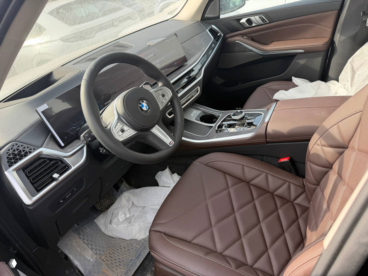 Вид салона водителя: рулевое колесо, цифровая панель и центральная консоль BMW X5, коричневая кожаная отделка и защитные чехлы на сиденьях.
