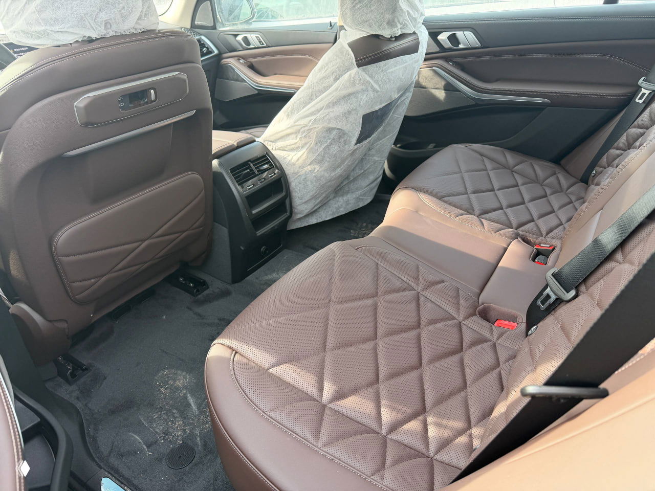 Задний ряд сидений BMW X5 2025 с коричневой стёганой кожей, видны напольные коврики и пространство для пассажиров с защитными чехлами.