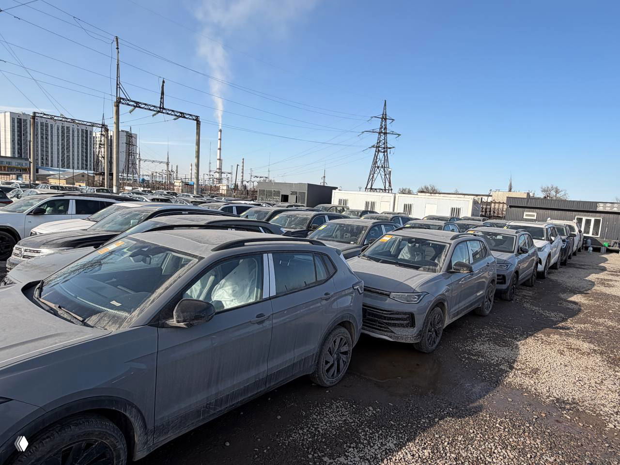 Общий план стоянки с множеством Volkswagen Tharu в ряд, контейнеры и городские здания на заднем плане, солнечный день, дорожная гравийная поверхность.