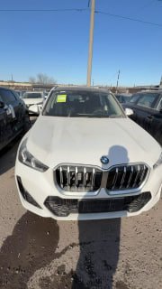 Передняя часть белого BMW X1 2025 на стоянке автосалона, видны решётка радиатора, фары, бампер и тень фотографа.