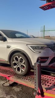 Крупный план переднего колеса белого кроссовера на рампе автовоза, вид спереди‑сбоку, видны опорные стойки и дорожная поверхность.