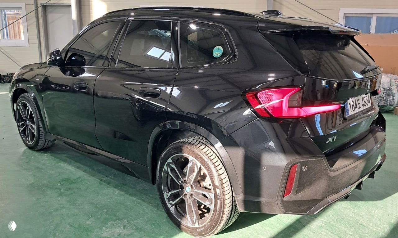 Боковой и задний вид чёрного BMW X1 в автосервисе: видна кормовая часть, световые элементы, колёса и кузов с отражениями освещения внутри помещения.