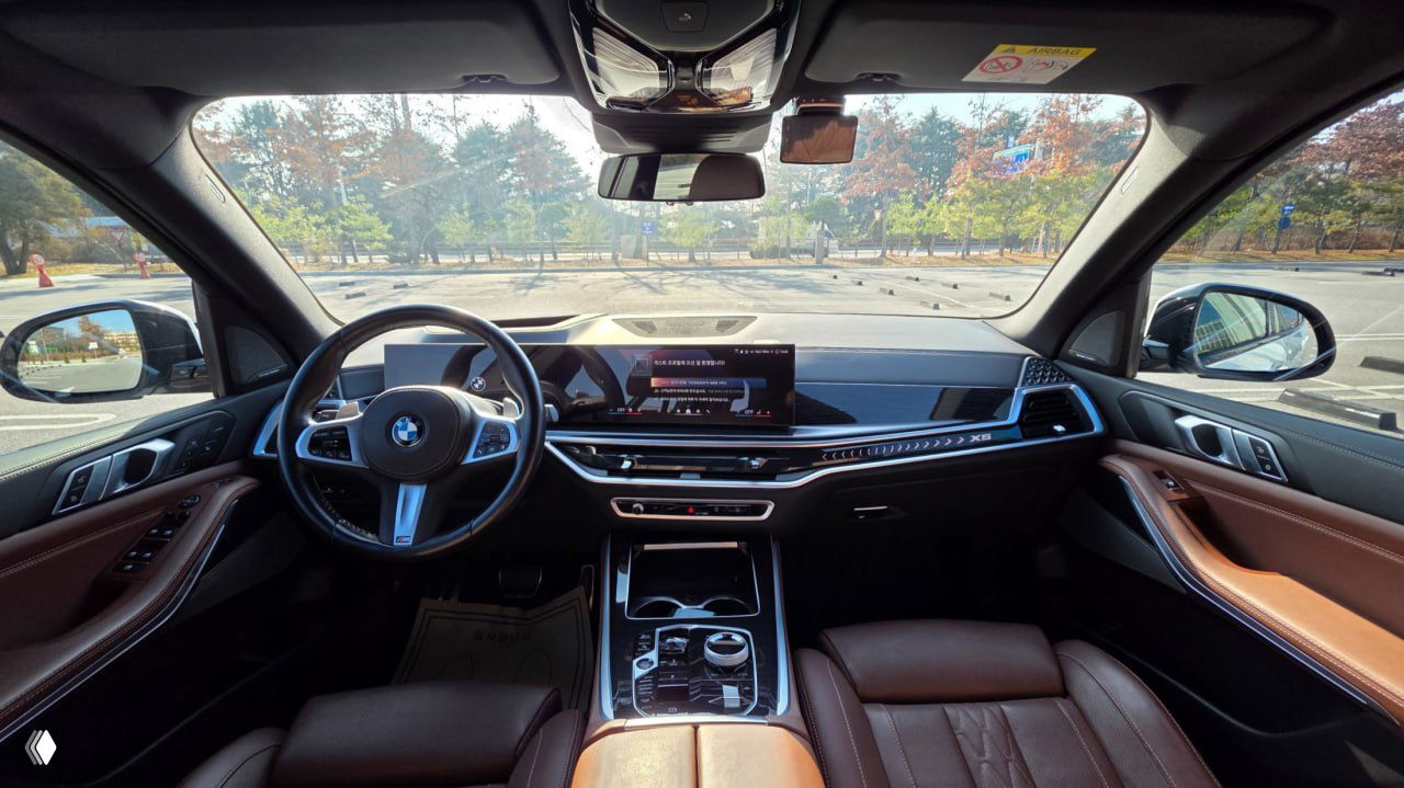 Салон BMW X5 xDrive 40i 2023 года — передняя панель, рулевое колесо, большой экран мультимедиа и коричневая кожаная отделка сидений.
