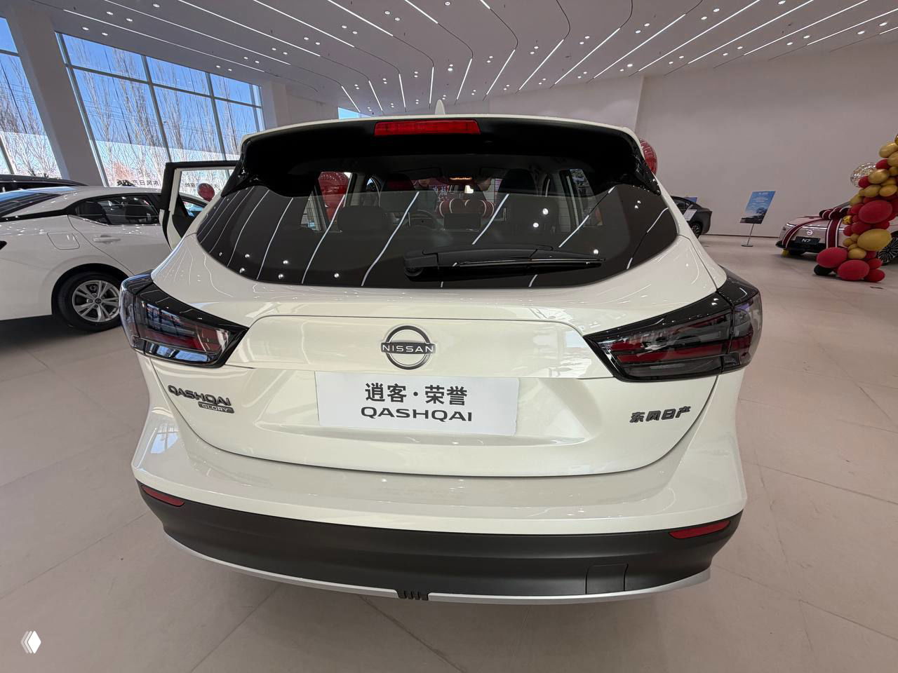 Вид сзади на белый Nissan Qashqai в салоне: эмблема, надпись QASHQAI и затемнённые фонари на светлом фоне шоу‑рума.