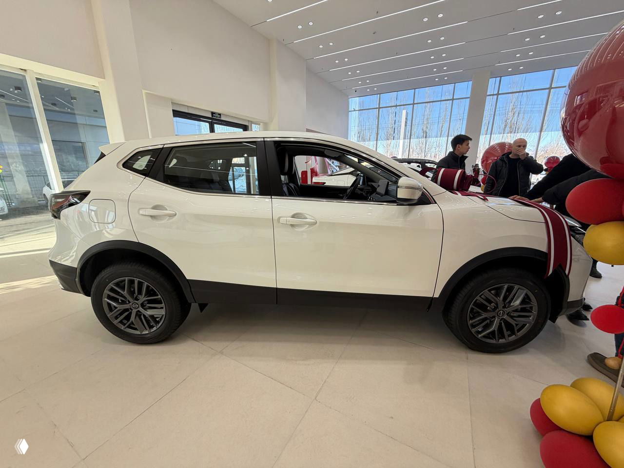 Боковой профиль белого Nissan Qashqai в салоне, видны колёса, кузов и просторное остекление на фоне больших панорамных окон.