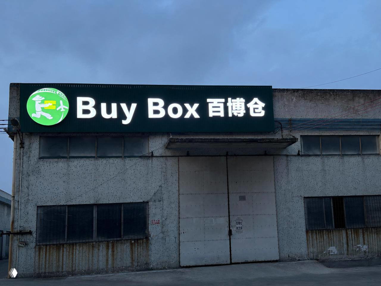 Фасад склада Buy Box в Китае в вечернее время: подсвеченная вывеска и закрытые промышленные ворота перед складским зданием.
