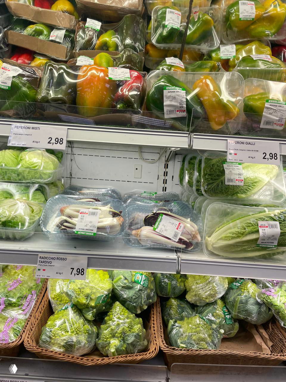 Полка овощей в итальянском супермаркете: запакованные болгарские перцы, радиккио, салаты и брокколи в корзинах, ценники видны.