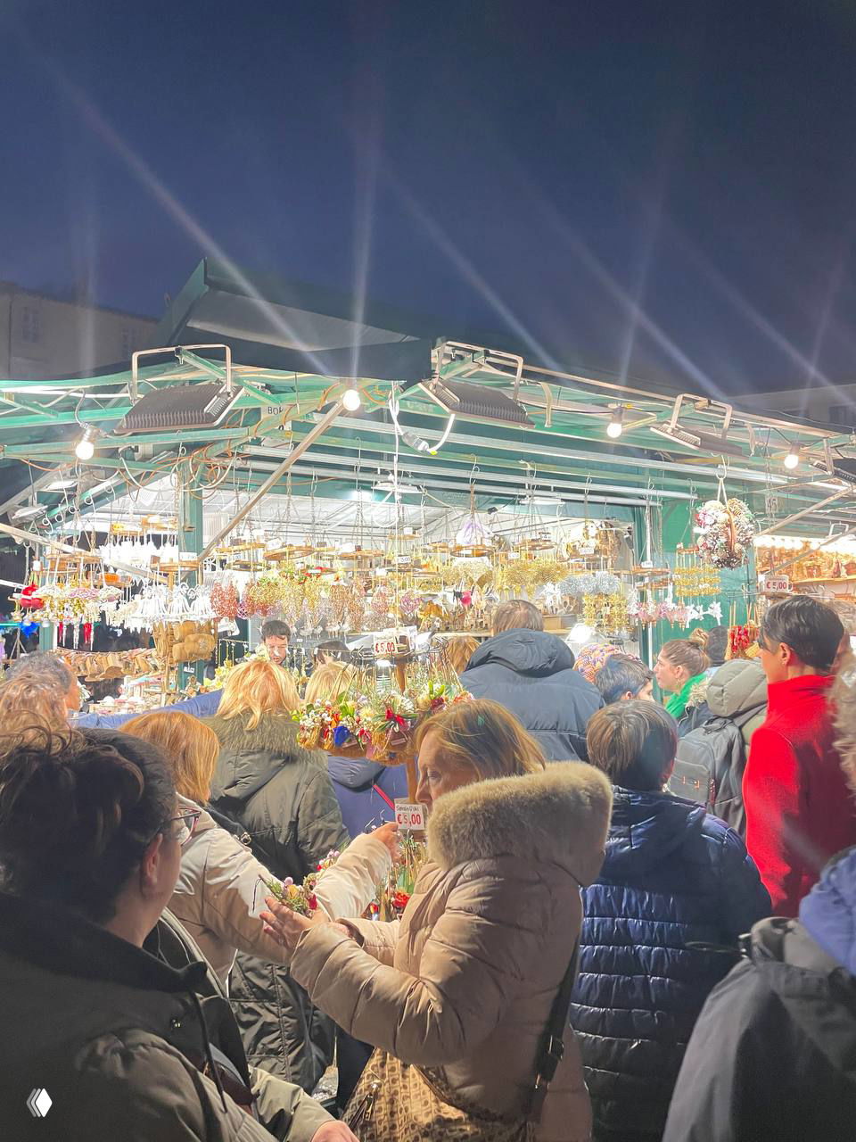 Плотная толпа людей у прилавков с рождественскими украшениями и гирляндами; ночная торговля на ярмарке в римской площади.