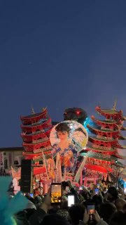 Вечерняя сцена карнавала: большой декоративный шар и элементы восточной архитектуры в декорациях, публика с телефонами и огни шоу.