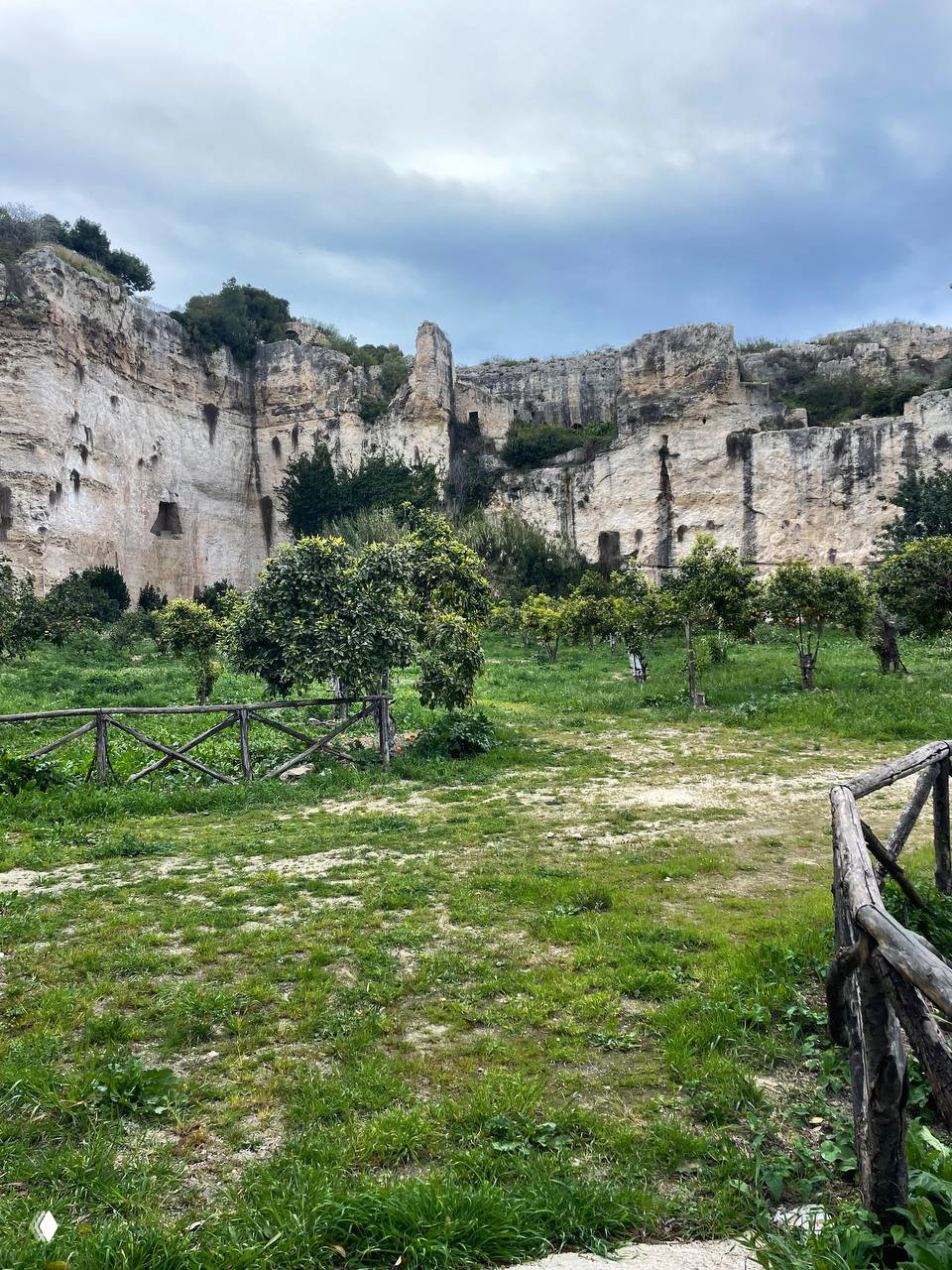 Скальные выступы и заросли с небольшой рощей или садом у подножия утёсов — вид напоминает карактерные каньоны и необычные природные ландшафты Сицилии.