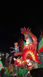 Артисты в пернатых ярких костюмах на огромной платформе, танцы и динамичная сцена на карнавале, многочисленная публика с поднятыми телефонами.