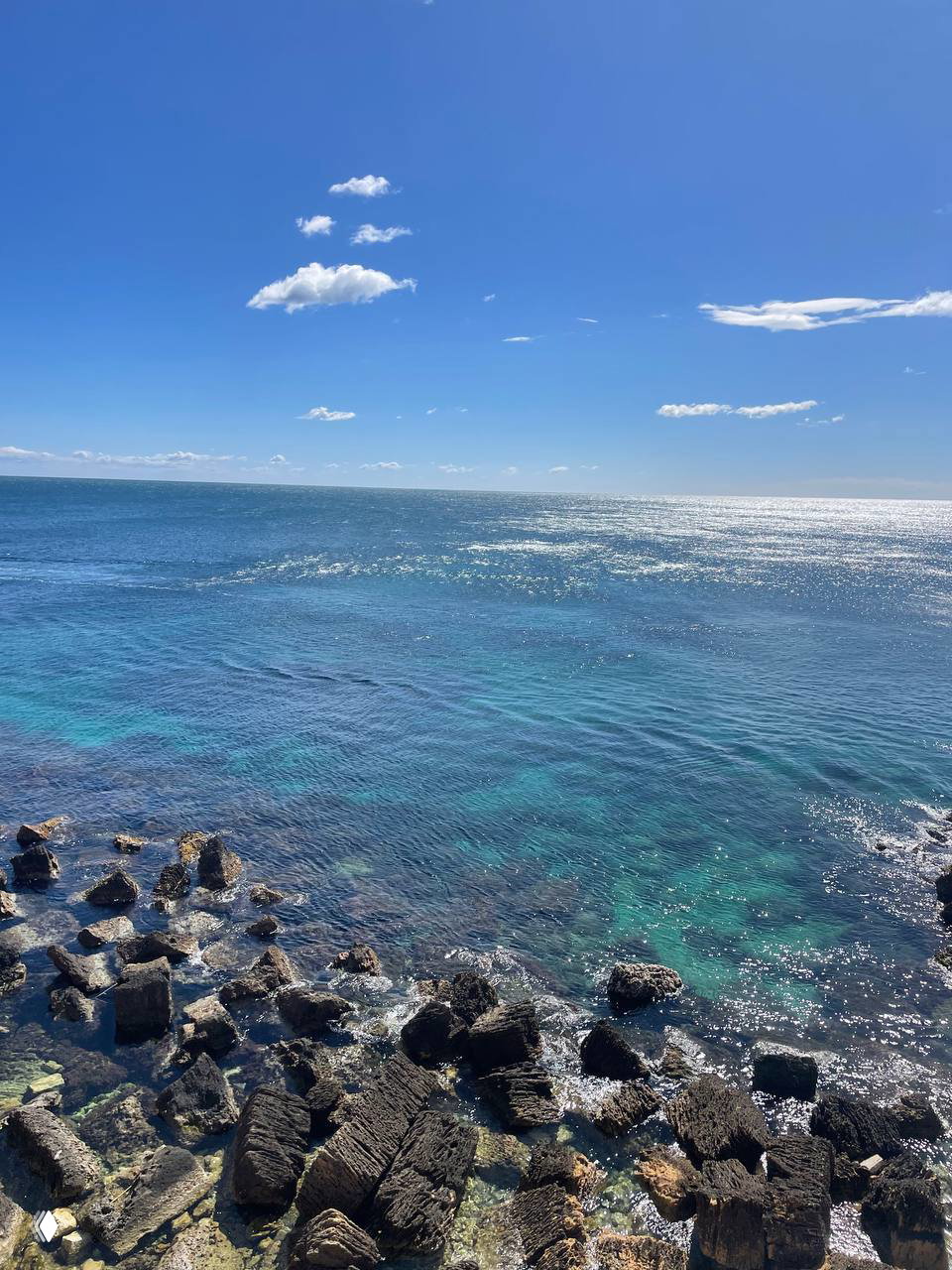Бирюзовое море Сиракузы с каменистым берегом и переливами на воде при ясном небе, вид от набережной — спокойная морская панорама.