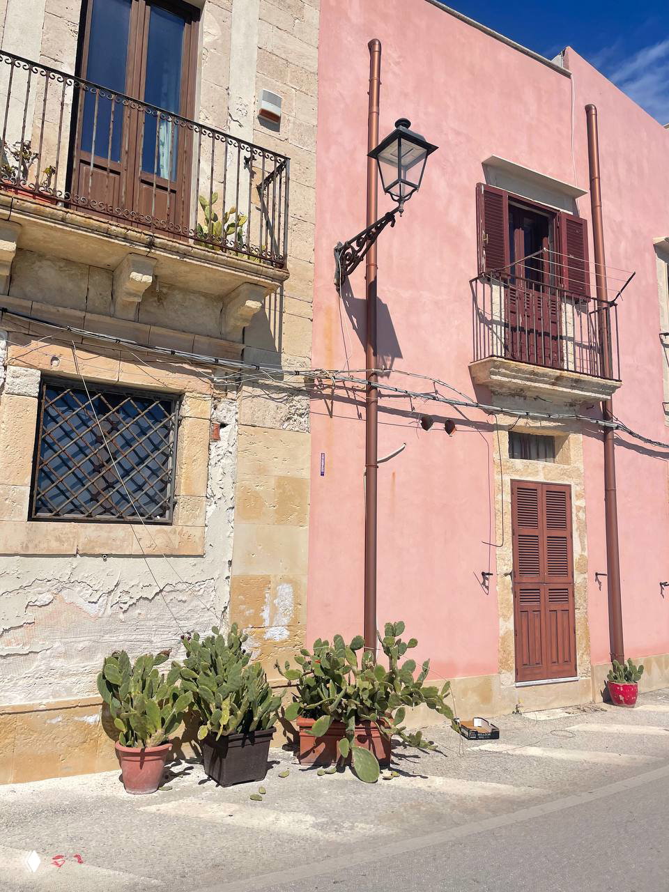 Уютный уличный уголок с розовым фасадом дома, балконом и горшками с кактусами на тротуаре — простая сценка сицилийского городка в солнечный день.