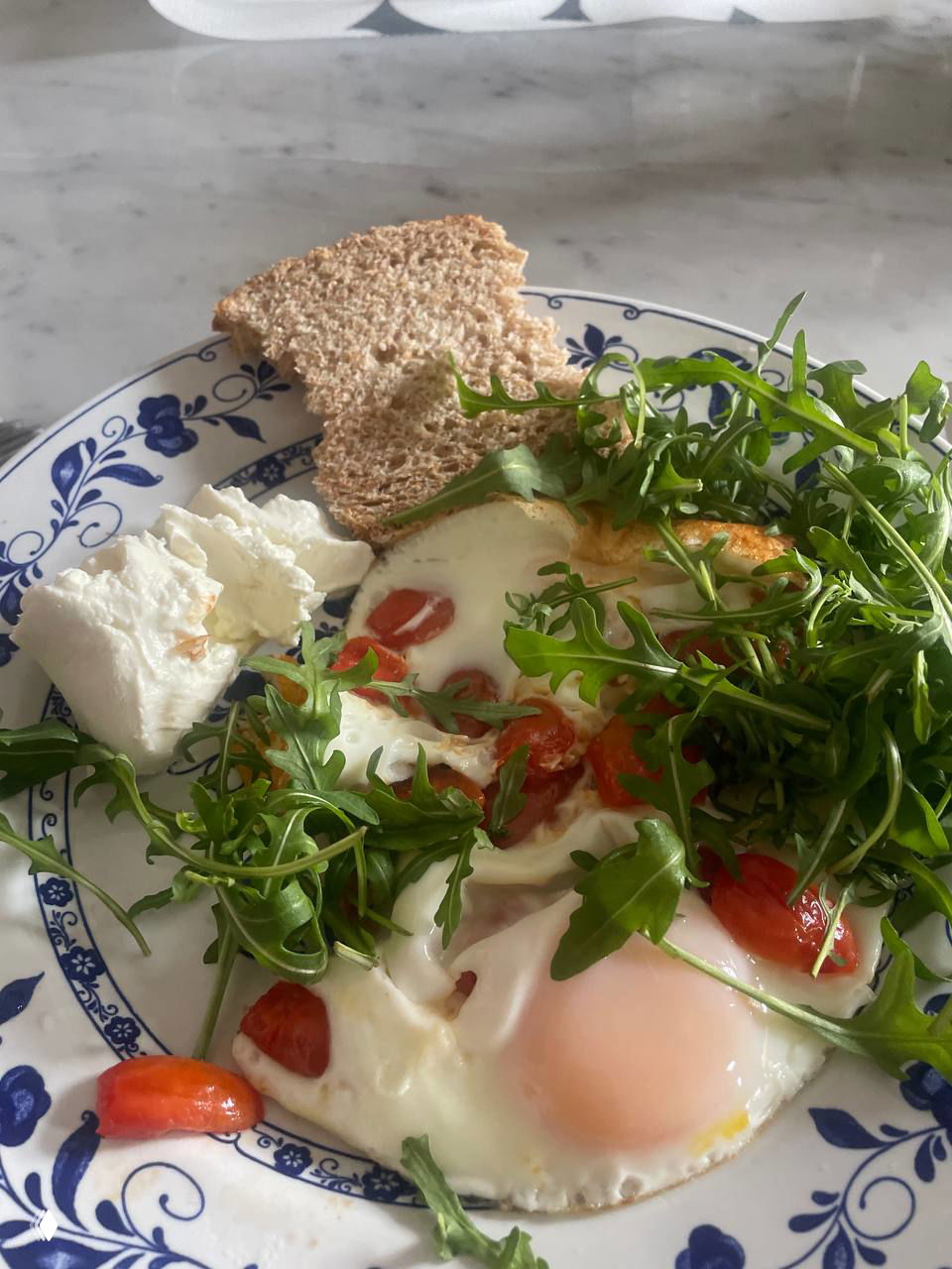 Тарелка с жареными яйцами, рукколой, кусочком хлеба и творогом — домашний утренний завтрак в крупном плане.