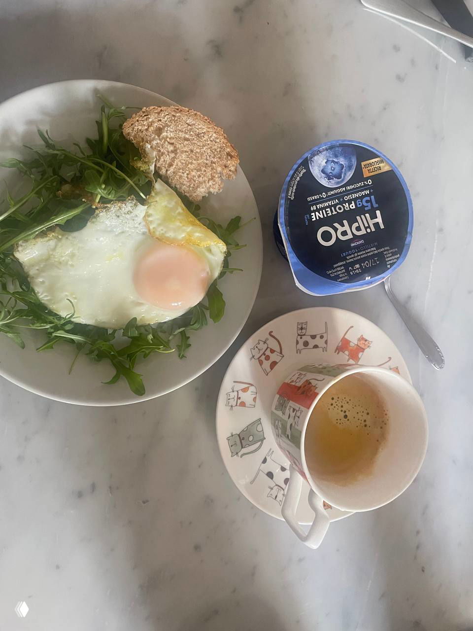 Вид сверху: тарелка с яйцом и рукколой, кусок хлеба, йогурт HiPRO и чашка кофе на светлом мраморном столе — утренний стол.