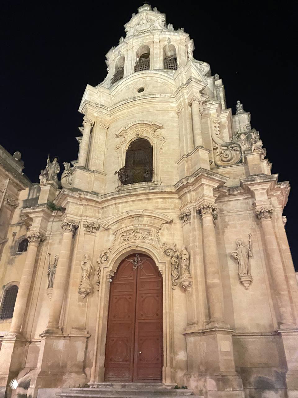 Крупный план барочного портала в Рагузе с резной дверью и лепниной; светлая каменная отделка и скульптурные детали на фоне ночного неба.