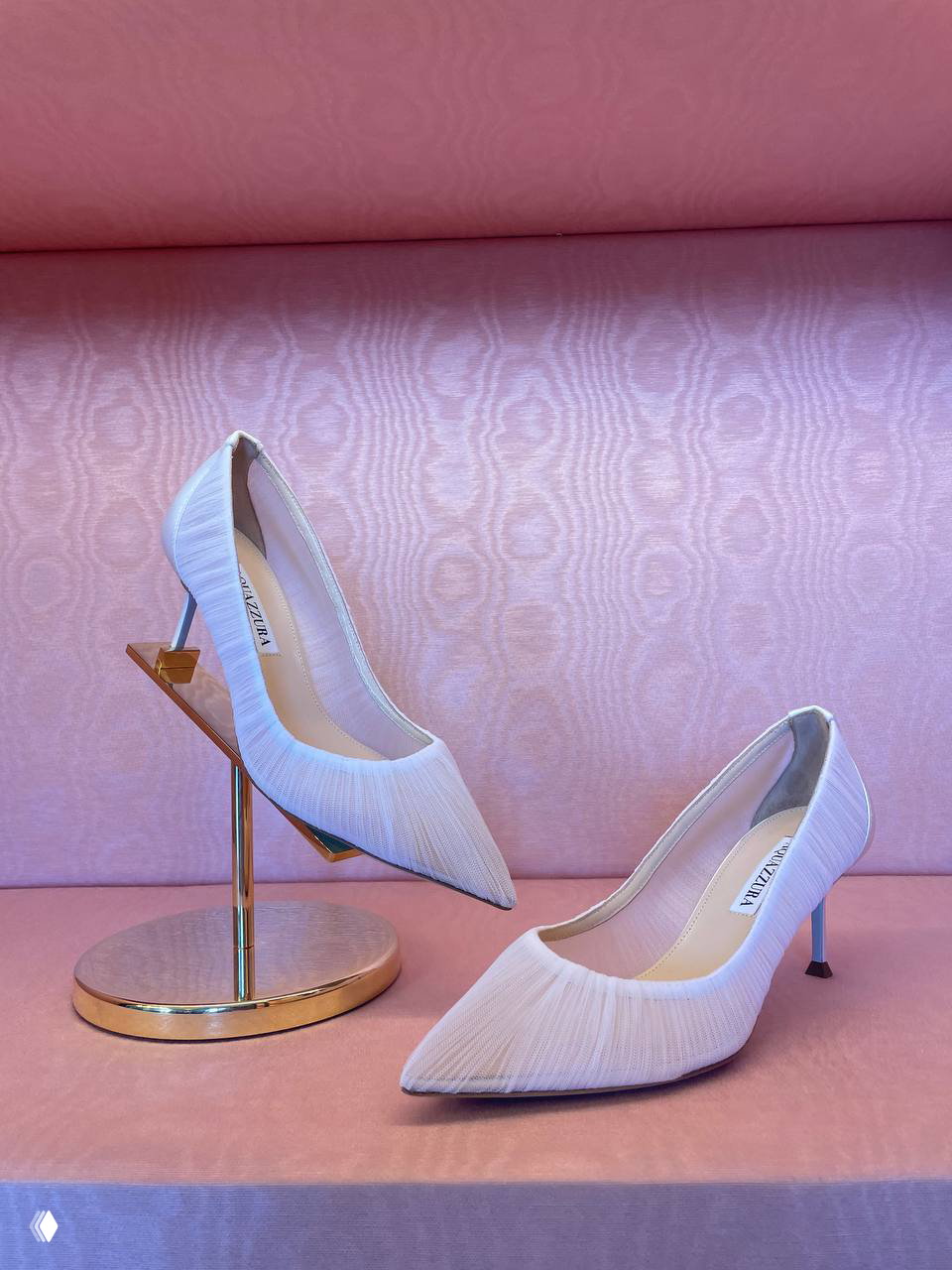 Витрина с белыми туфлями Aquazzura на розовом фоне: минималистичные подставки, текстурная обивка и акцент на форме обуви.