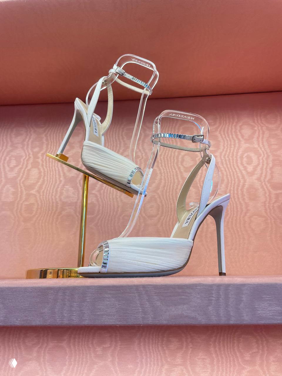 Пара высоких босоножек Aquazzura на золотистой подставке и розовой полке: акцент на деталях ремешков и декоре каблука в бутике.