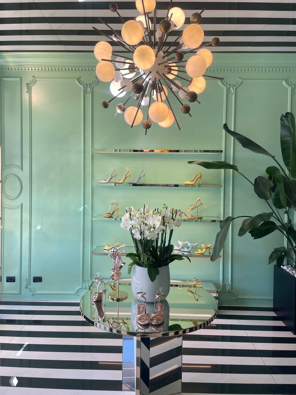 Интерьер Aquazzura с мятно-зелёной стеной, зеркальным столом и орхидеями на переднем плане; на полках — туфли и босоножки бренда.